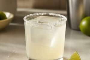 Margarita pâle dans un verre bas au bord salé, gros glaçon et quartier de citron vert, sans marque ni personne