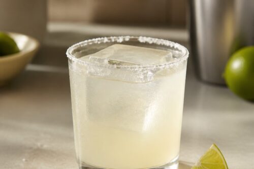 Margarita pâle dans un verre bas au bord salé, gros glaçon et quartier de citron vert, sans marque ni personne