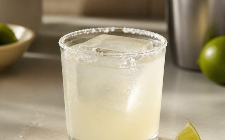 Margarita pâle dans un verre bas au bord salé, gros glaçon et quartier de citron vert, sans marque ni personne